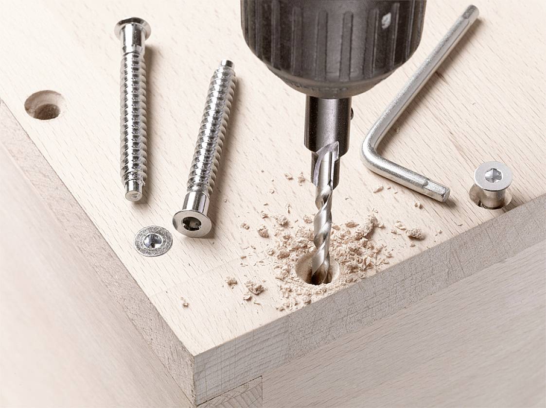 Wooden board with a drill, drilling a hole. Wood shavings around the hole. Screws and an Allen key are lying beside it.