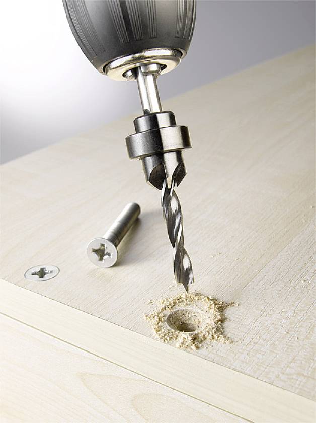 A drill creates a hole in a wooden beam. A screw lies beside the hole. Wood shavings are visible around the hole.