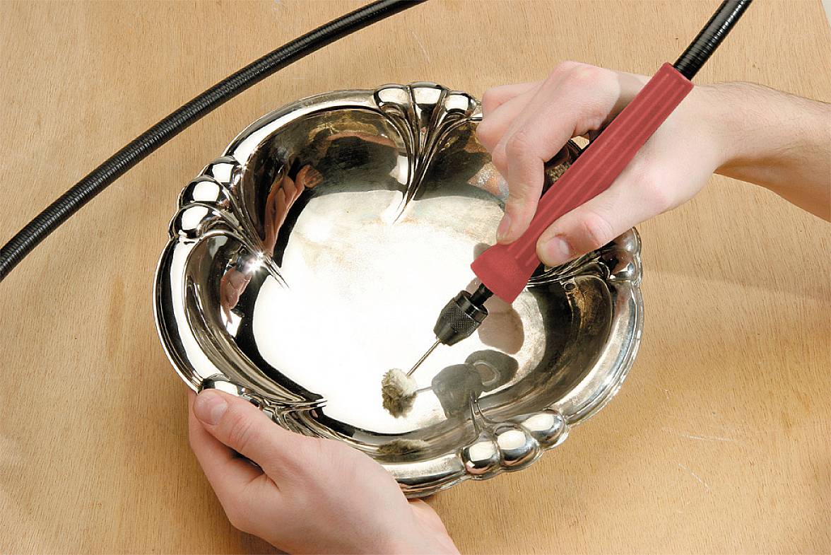 A person is polishing a silver, decorative plate with a red and black tool on a wooden table.