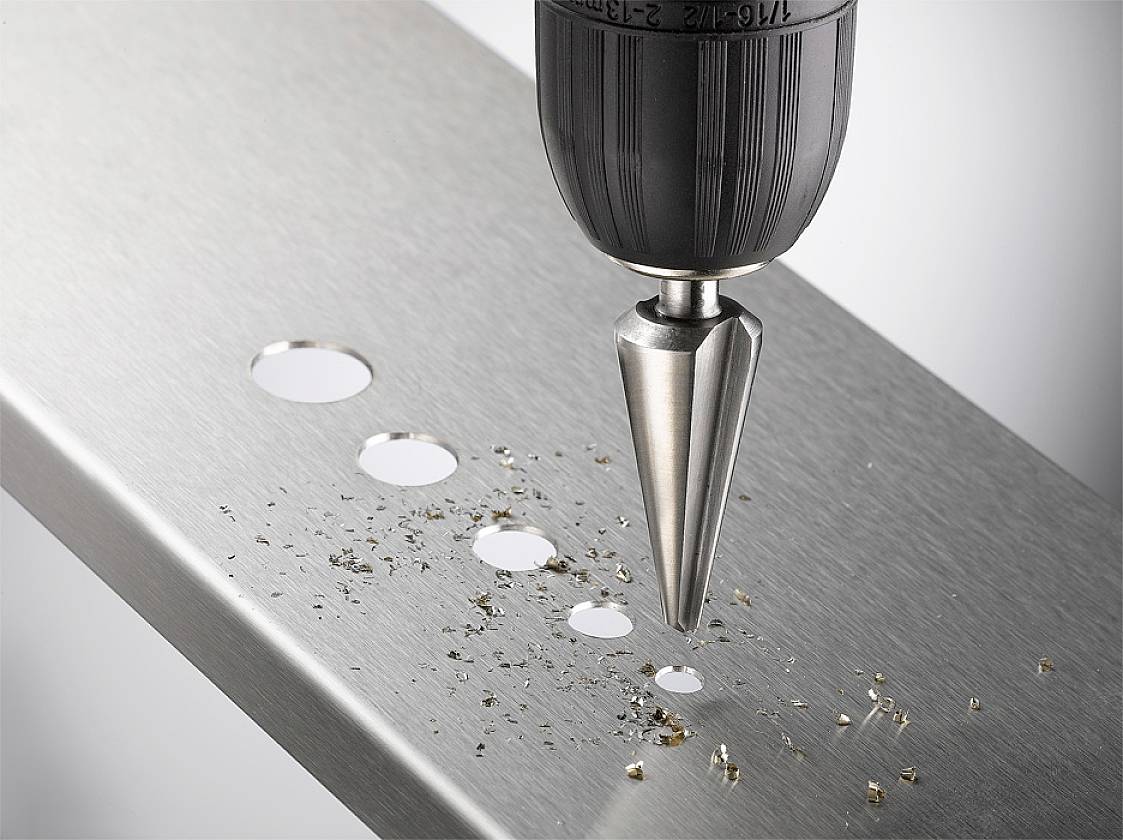 A tapered drill makes several holes of different sizes in a metal plate. Metal shavings are scattered around the holes.