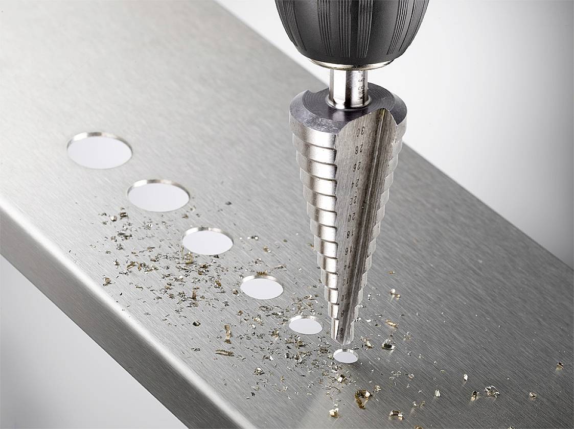 A stepped drill bit is drilling a hole in a metal plate. Several drilled holes of varying sizes are visible, with metal shavings lying nearby.