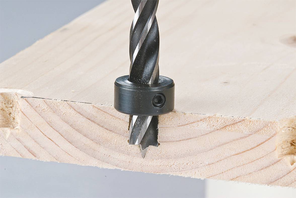 A black drill bit is piercing a piece of wood. A stop ring is attached to the drill bit to control the depth of the hole.