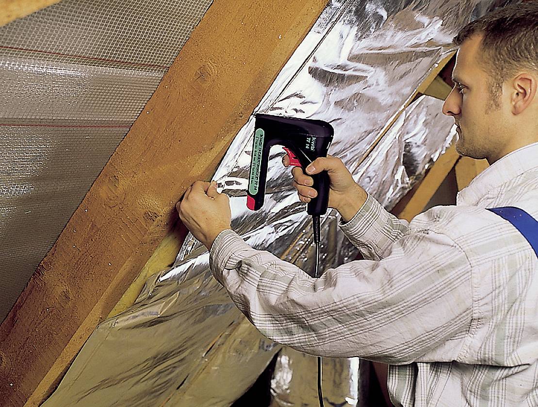 A man is securing insulation with a hot air gun in an attic. The insulation consists of silver foil.
