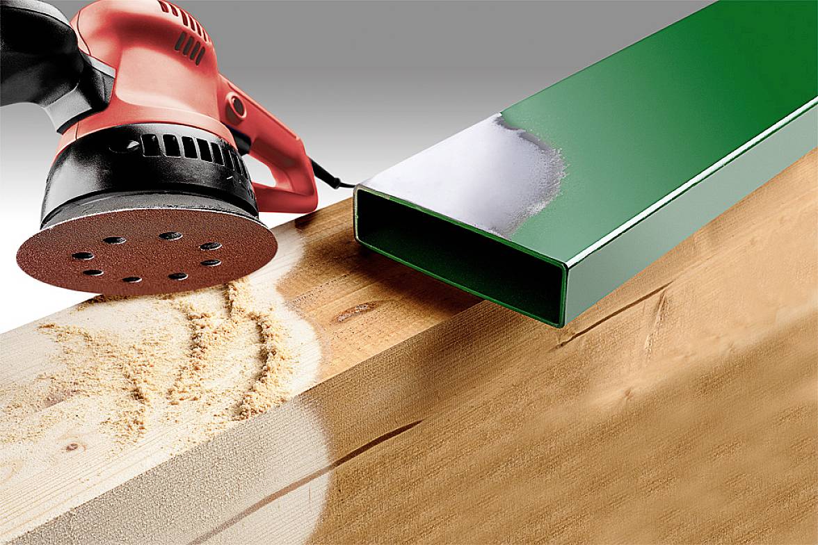 An electric sander is working on a wooden board, which lies next to a green metal profile. Dust is visible on the wood.