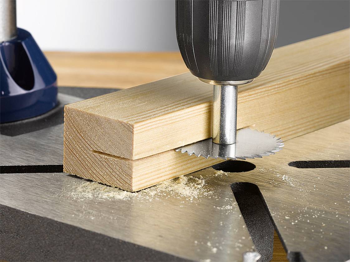 A drill with a hole saw attachment is drilling a round hole in a wooden board. Sawdust is scattered around the spot.