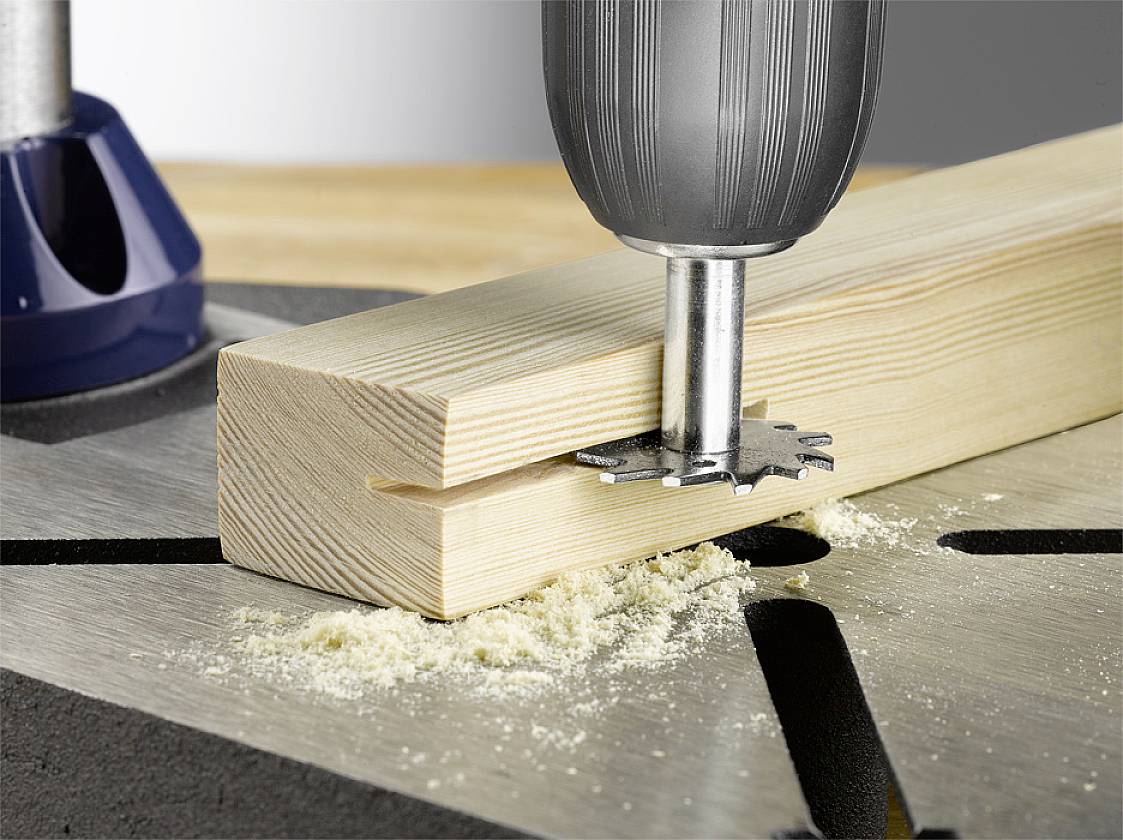 A drill bit is machining a piece of wood, drilling a hole. Wood shavings are visible.