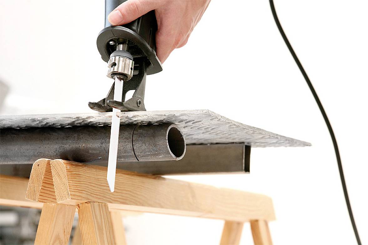 A hand is holding an electric saw cutting through a metal pipe. The pipe is resting on a wooden scaffolding.