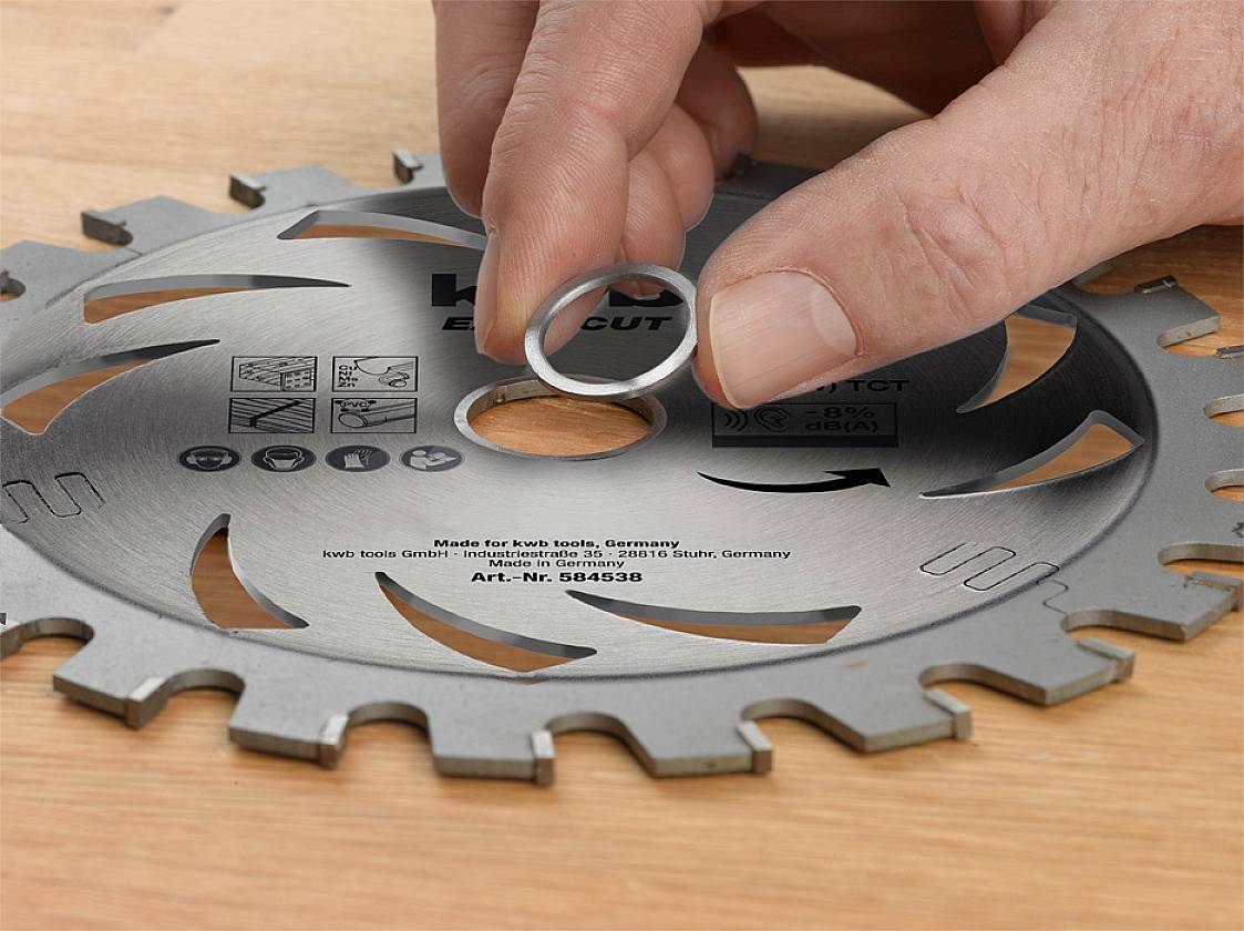 A hand places a metal ring onto a circular saw blade's mounting hole to adjust the diameter. The blade is resting on a wooden surface.