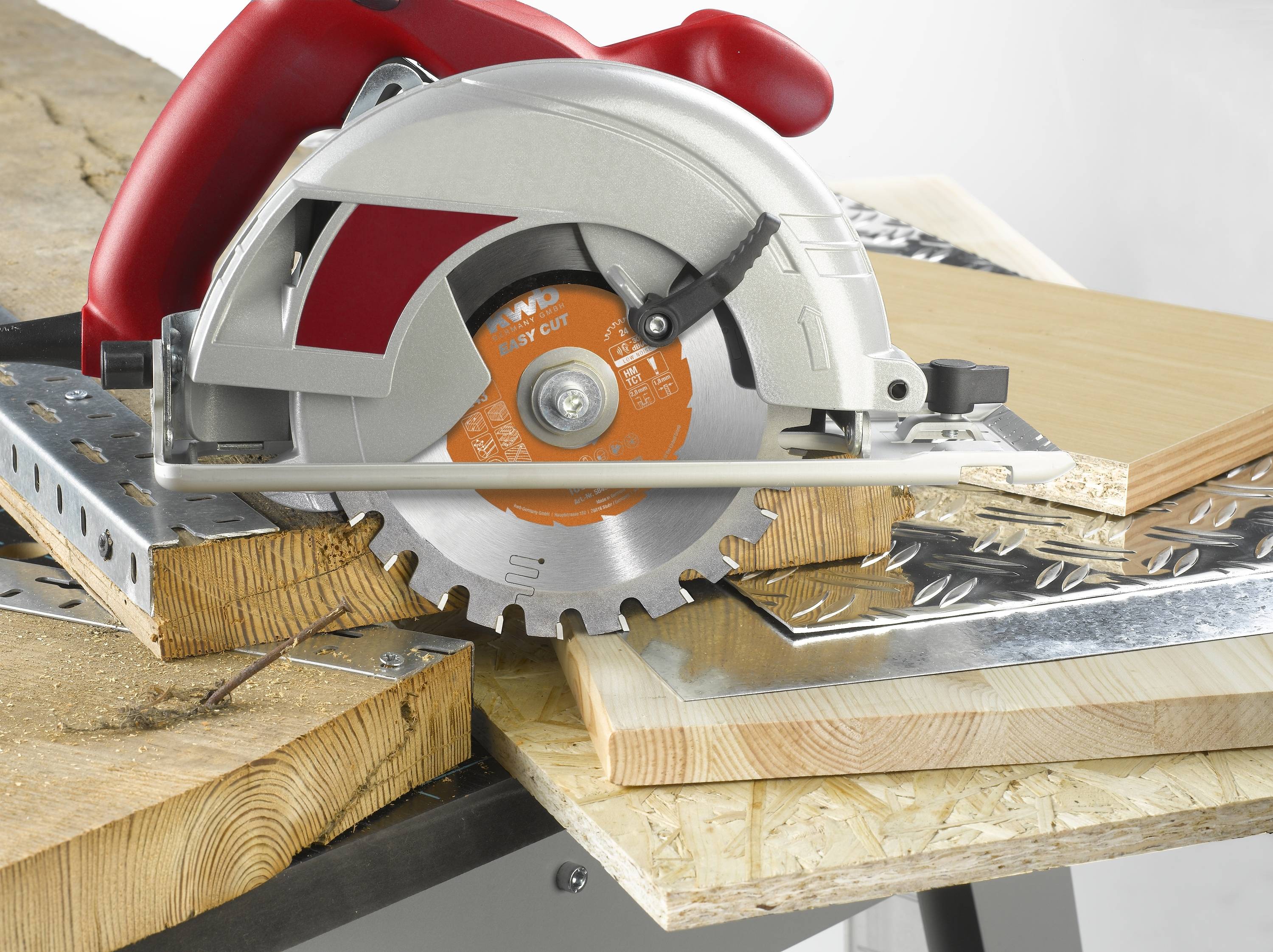 A circular saw cuts through several wooden and metal plates on a workbench. Various types of materials are visible.