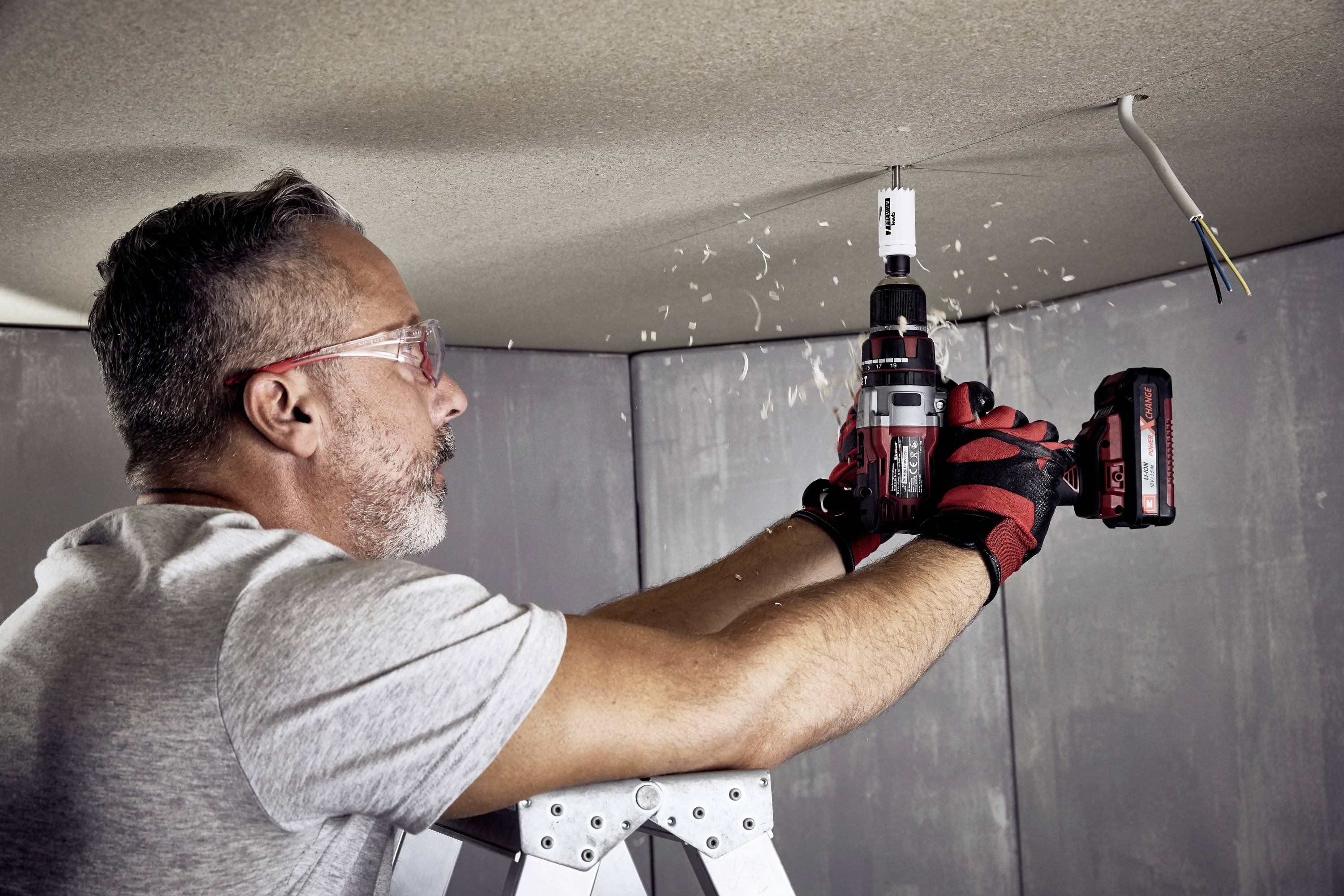 A man on a ladder is drilling a hole in the ceiling. He is wearing safety glasses and gloves to ensure safety.
