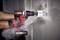 A person is drilling a hole in a wall with a cordless drill. Dust is spreading around the drilling area. Electrical cables are visible.