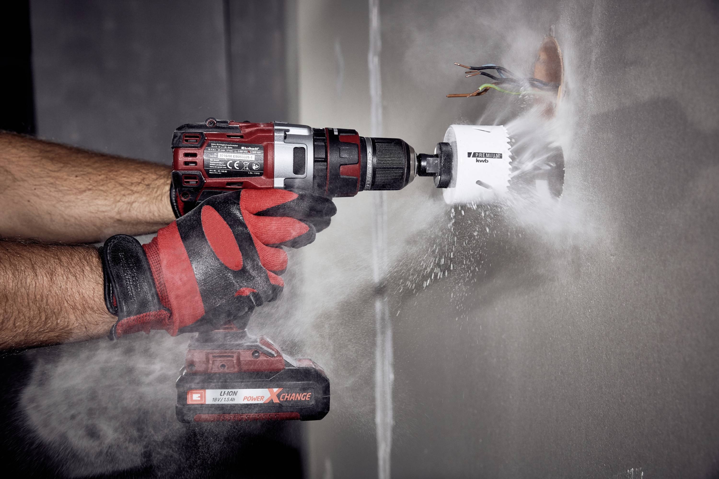 A person is drilling a hole in a wall with a red cordless drill. Dust is flying into the air. Several cables are visible in the hole.