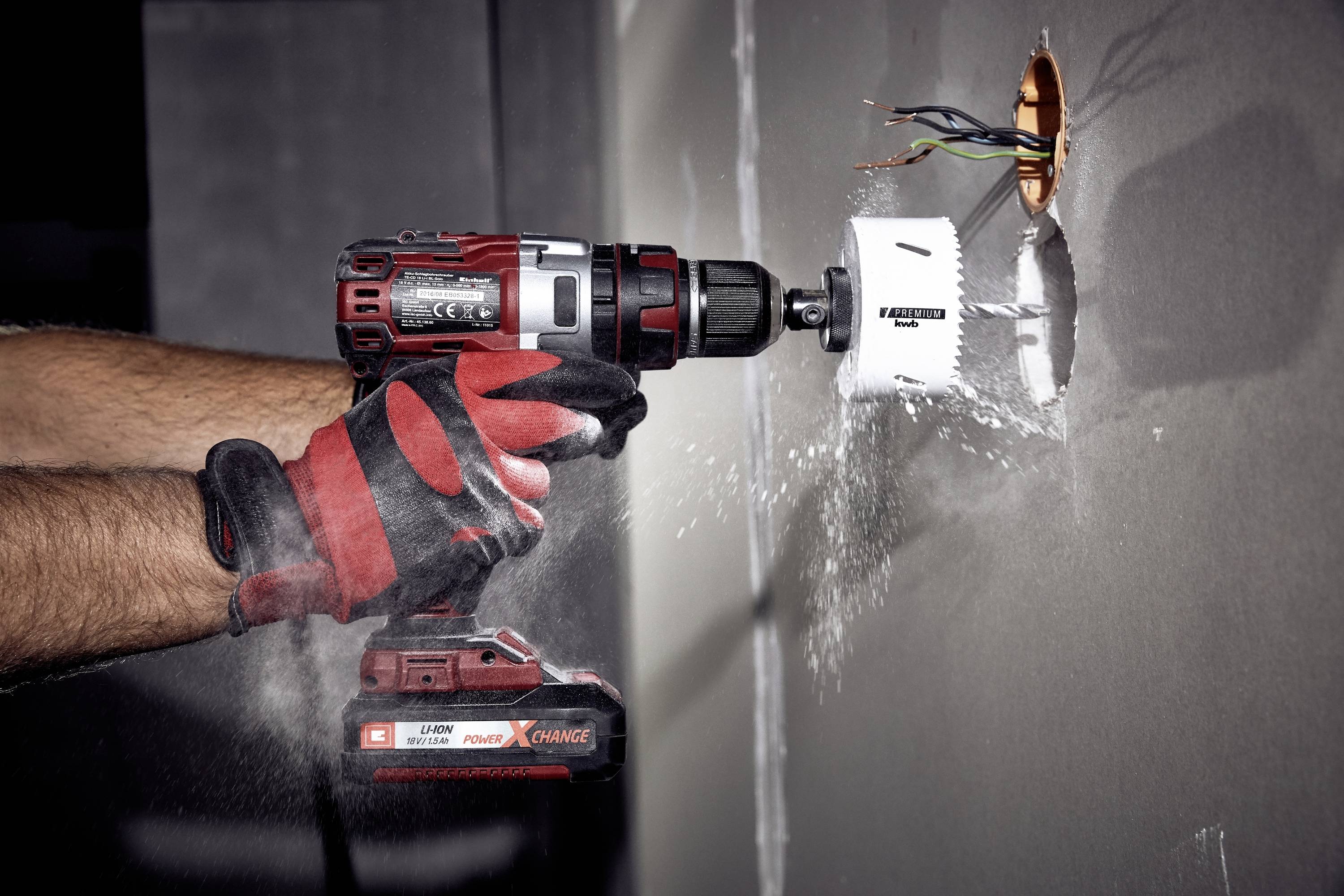A person is wearing red gloves and using an electric drill to bore a round hole in a wall.