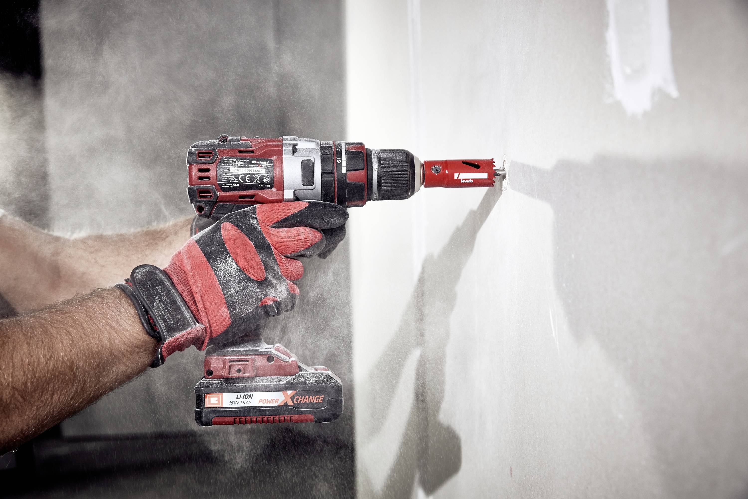 A person is drilling a hole in the white wall using a red cordless drill. Dust is flying around.