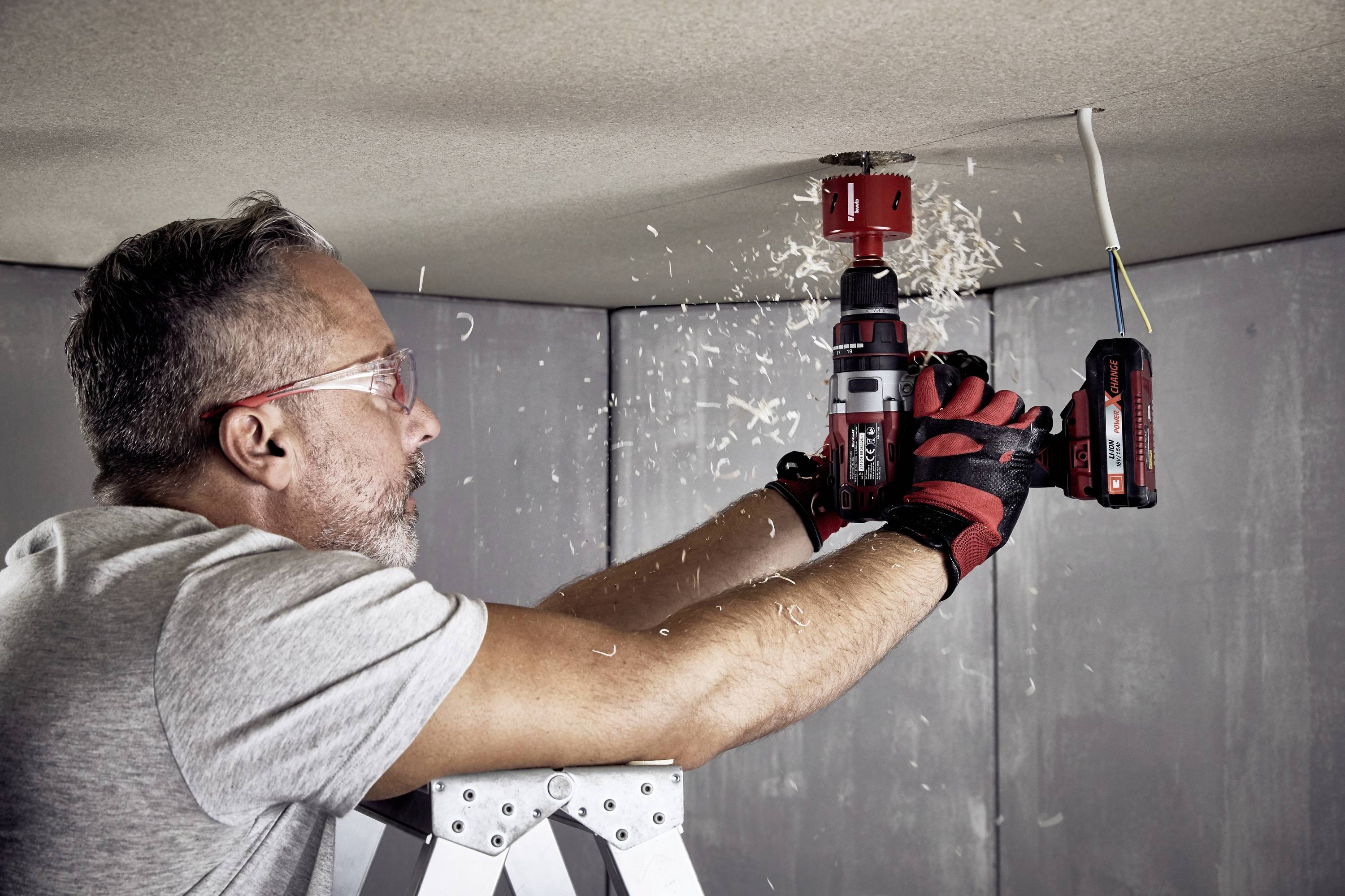 A man on a ladder is drilling a hole in the ceiling with a red drill. Wood shavings are falling down, and he is wearing safety glasses and gloves.