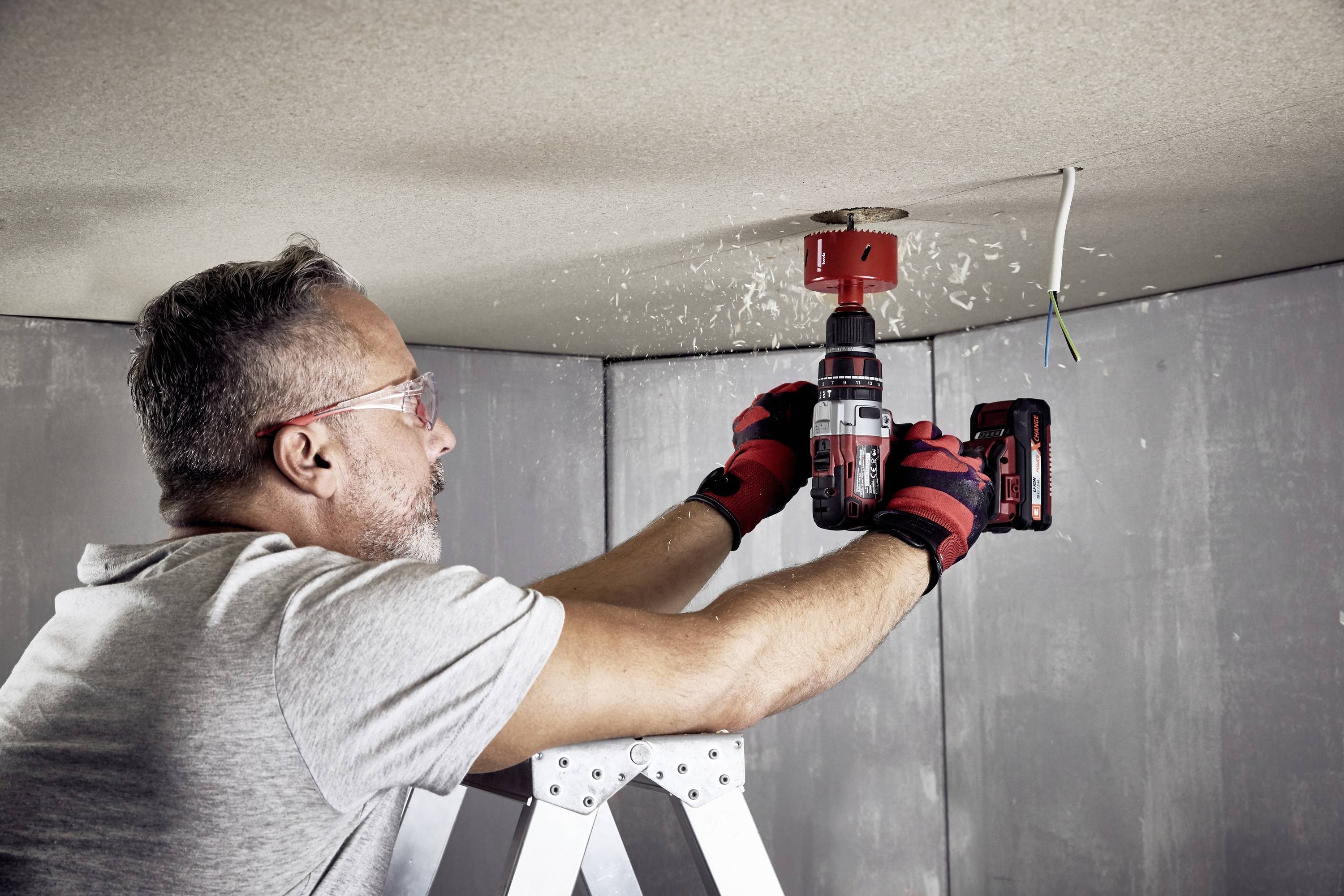 A man on a ladder is drilling a hole in a ceiling with a cordless drill, with wood shavings falling down.