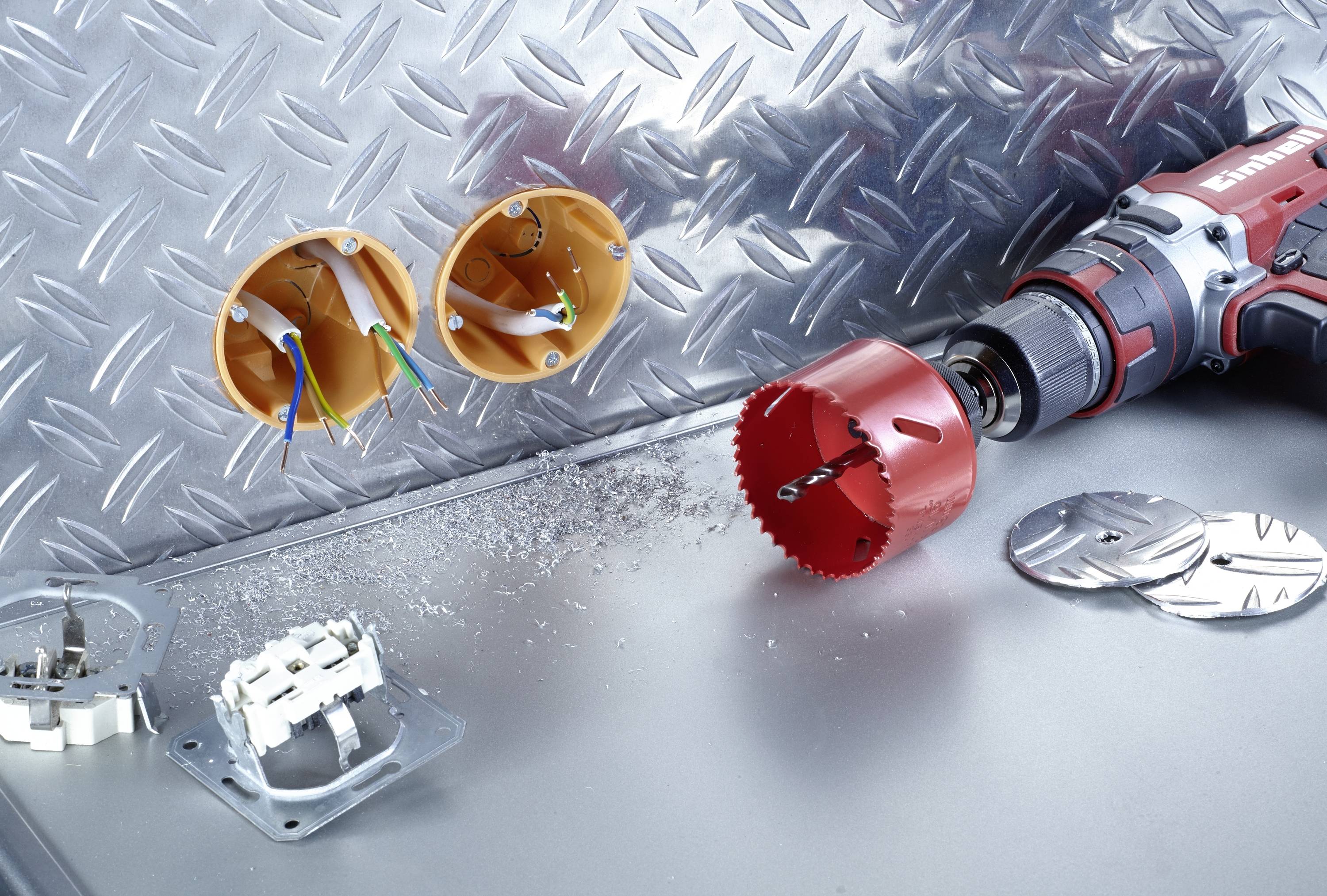 A red drilling tool with a hole saw head is lying on a metal surface. Beside it are two empty orange socket boxes with cables.