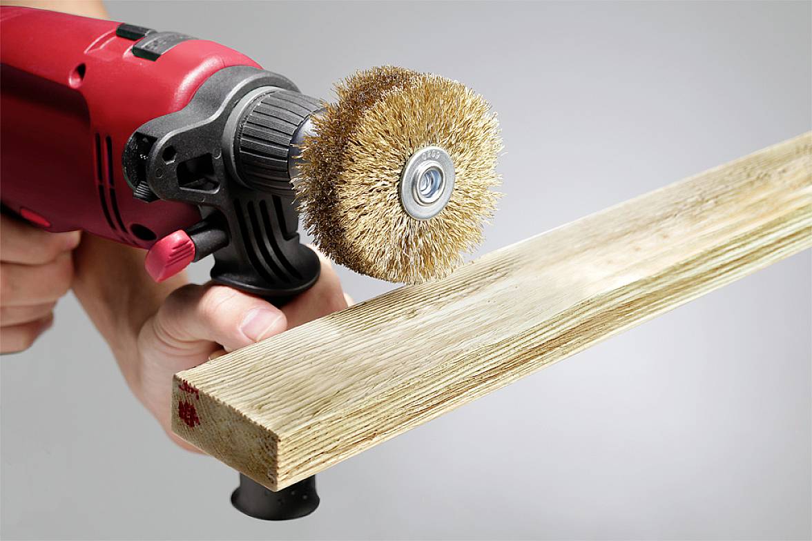 'A hand is holding a red and black drill with a wire brush attachment, which is grinding a wooden board.'