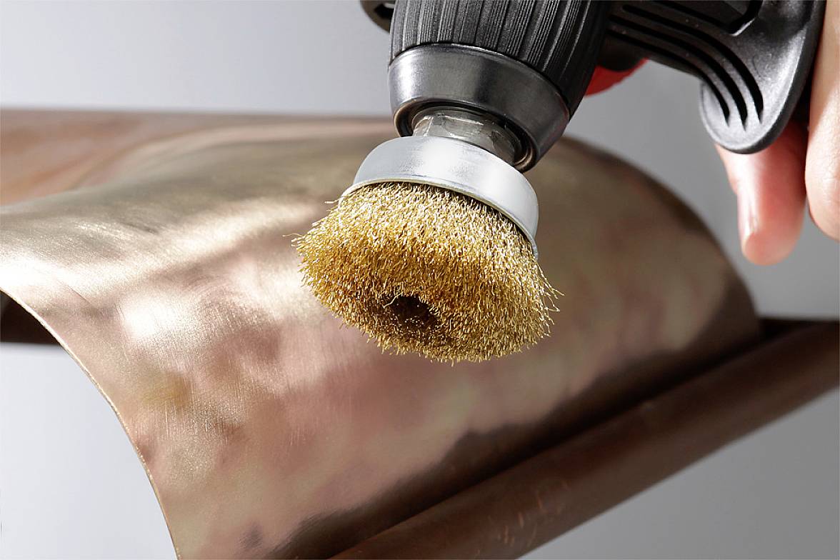 A wire brush is polishing a metal surface, possibly copper, for cleaning or surface treatment.