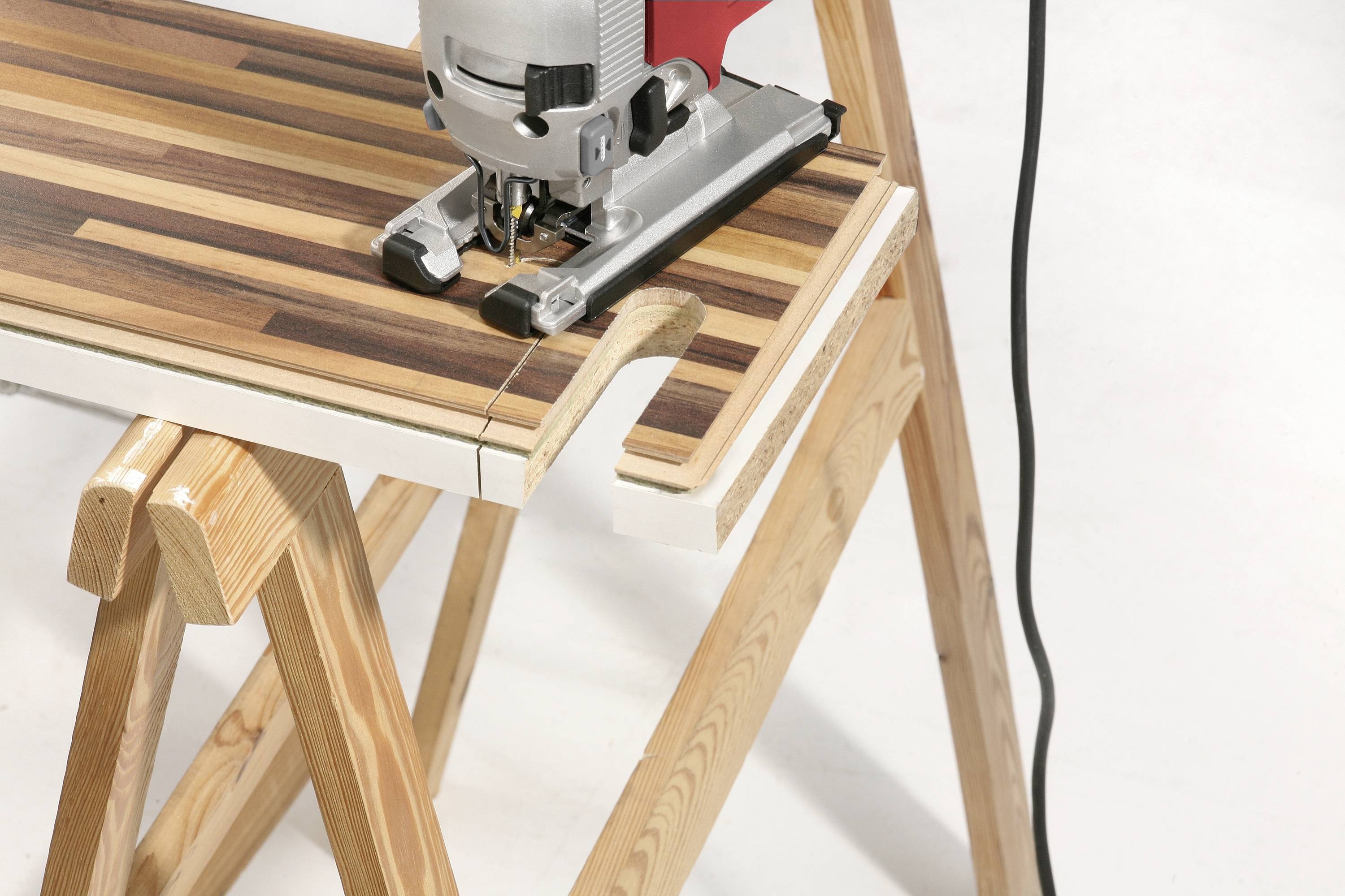 A jigsaw is cutting a wooden board on two sawhorses. The wood has a striped appearance in various shades of brown.