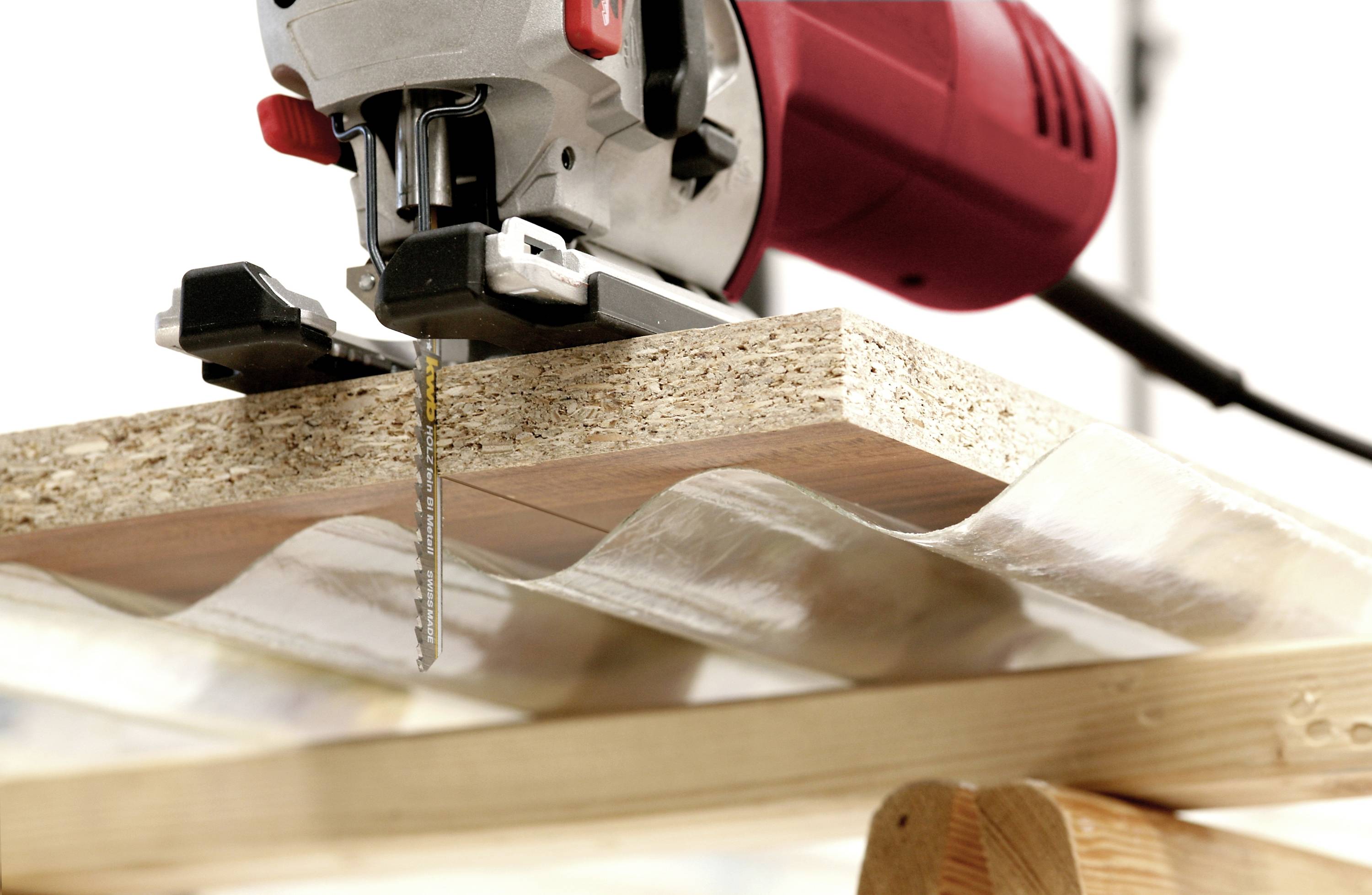 An electric jigsaw cuts through a compressed fibreboard board lying on two trestles, with a red handle and sharp saw teeth.