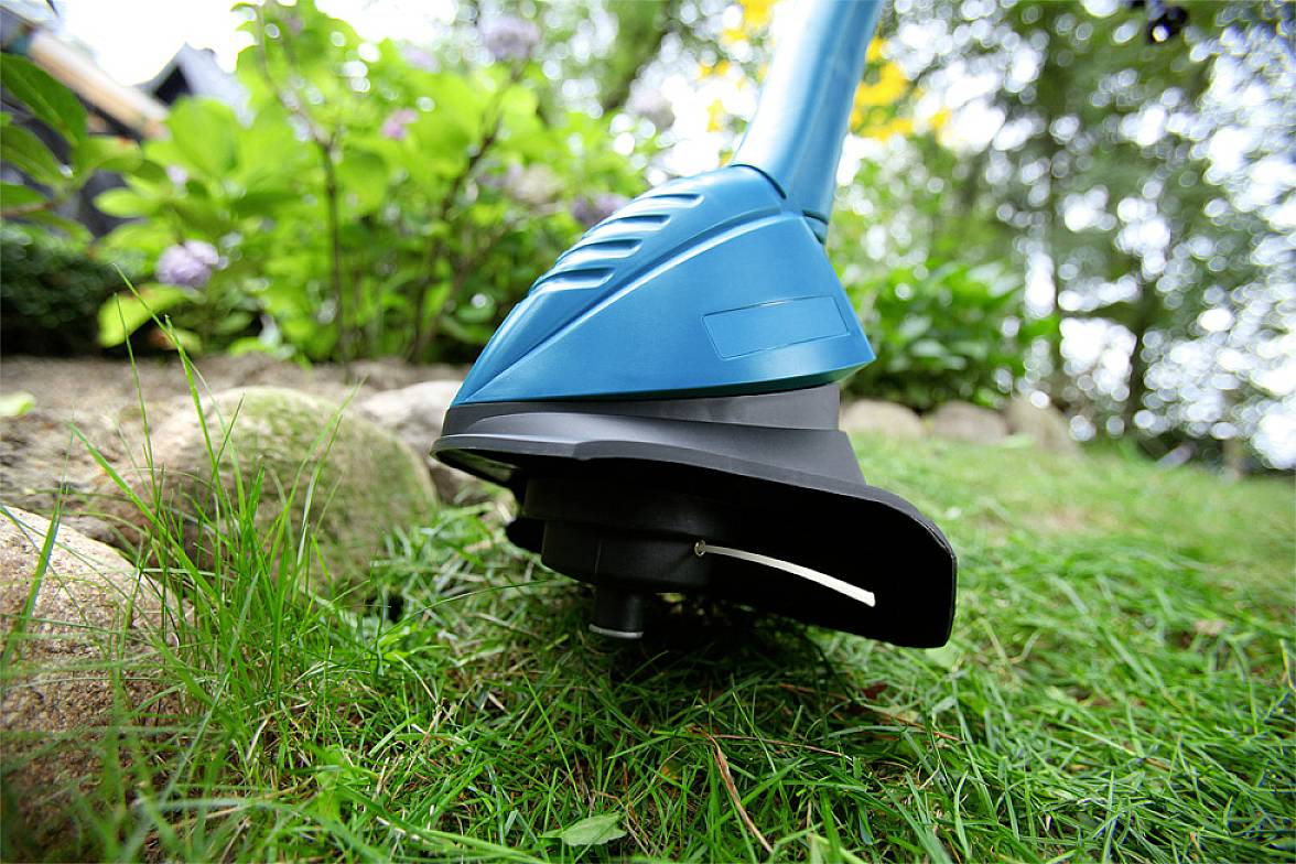 A blue lawn trimmer is cutting grass at the edge of a garden. Plants and trees can be seen in the background.