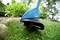 A blue lawn trimmer is cutting grass at the edge of a garden. Plants and trees can be seen in the background.
