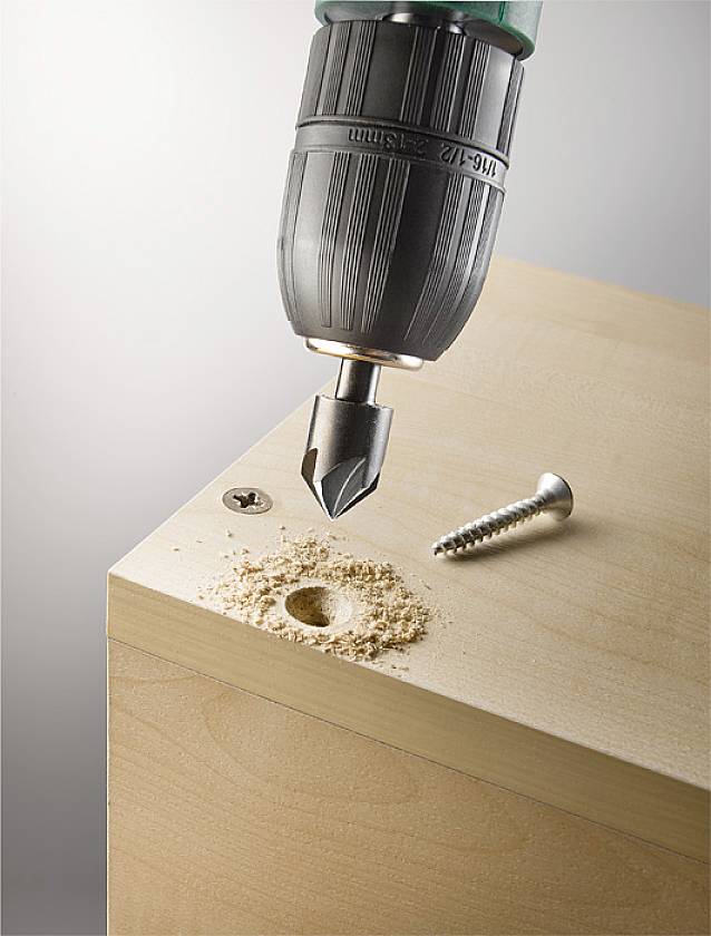 A drill is boring a hole into a wooden board, next to an already screwed-in screw. Wood shavings are scattered around the hole.