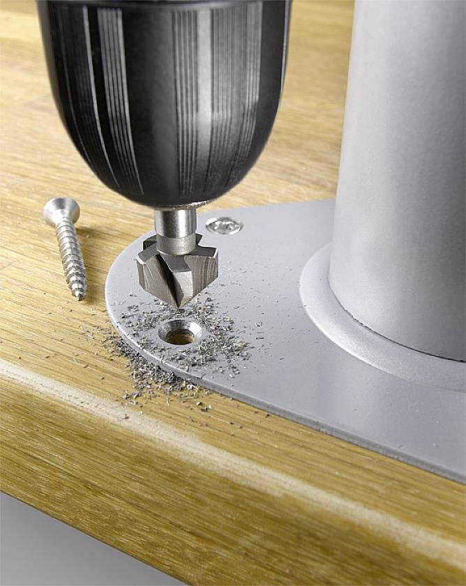 A drill is lowering a screw into a metal plate mounted on wood. Another screw lies beside it.