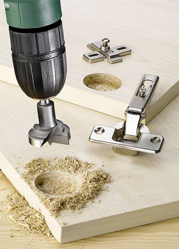 A green drill with a Forstner bit is precisely drilling holes in wooden boards, with a hinge mounted on it.