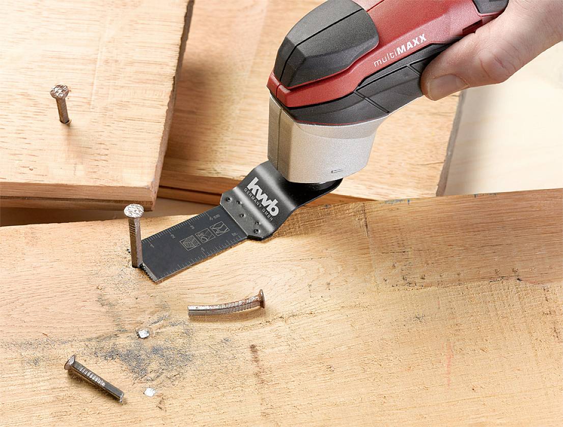 A hand is holding a multi-tool that is currently cutting nails through wood. Cut pieces of the nails are already lying on the wood.
