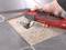 A hand is holding an oscillating multi-tool that is removing grout from between grey tiles.