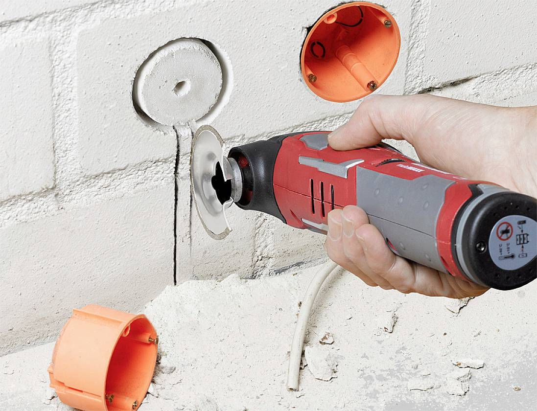 A person is using a red and grey tool to cut holes in a white brick wall, with visible orange electrical back boxes.