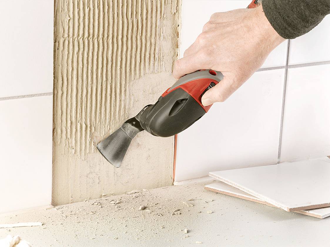 'Hand holding an electric tool', removing applications from a wall. Dust and ceramic tiles are visible.