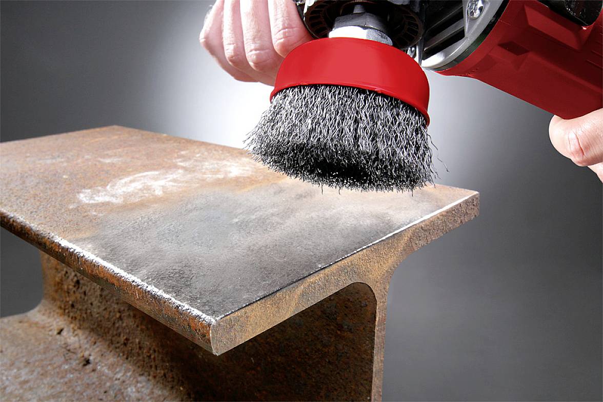 A wire brush removes rust from a metal carrier. Hands hold the tool securely. The carrier shows clear signs of corrosion.