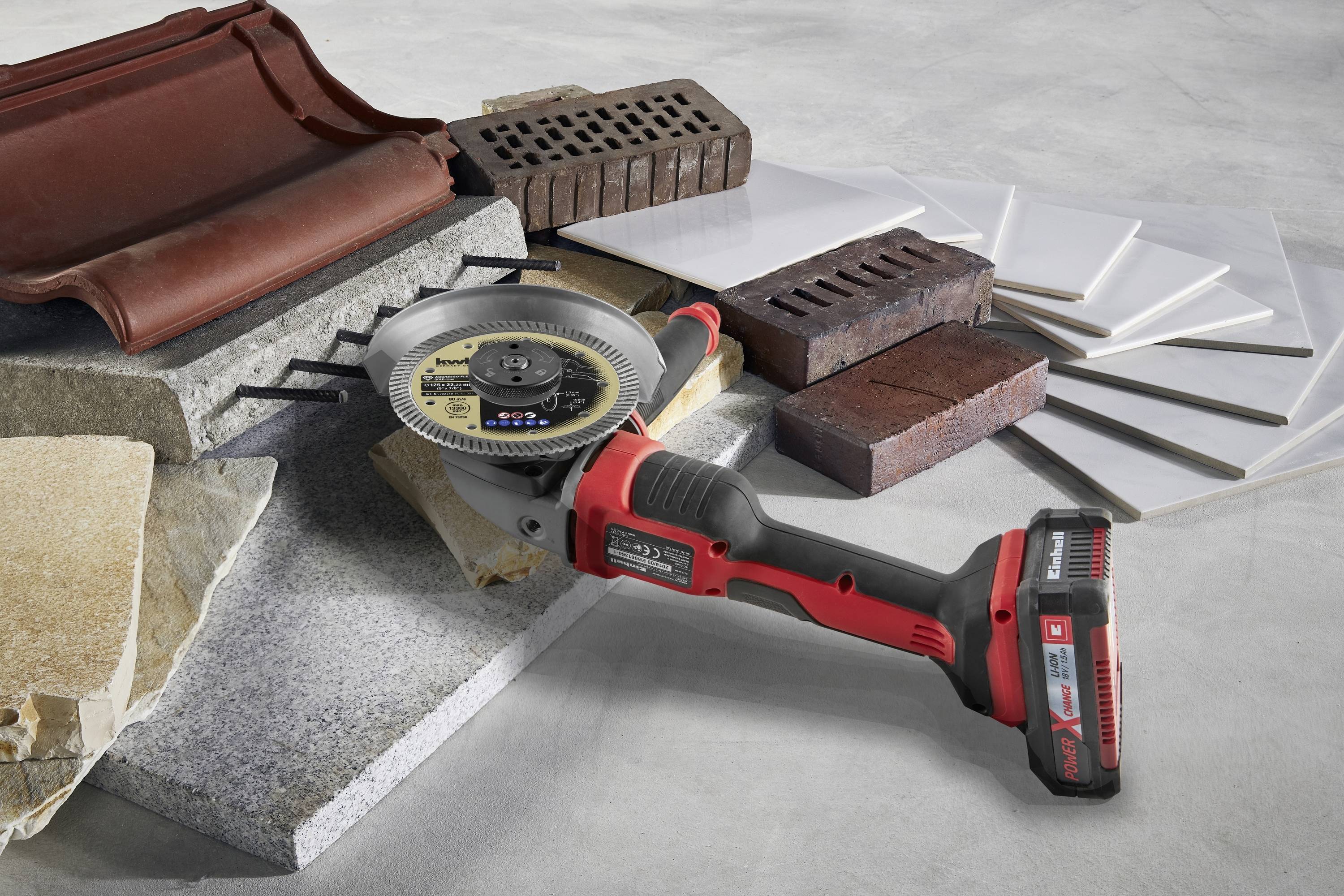 A red cordless power tool is resting on various building materials such as tiles, bricks, and roof tiles on a concrete surface.