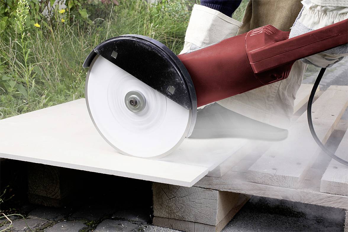 A worker is using a red electric cutting disc to cut a large white board outdoors. Dust is being created.
