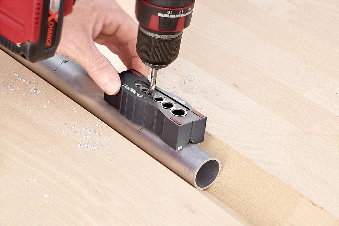 A person is drilling holes in a metal pipe using an electric drill, with a drilling template used for guidance.