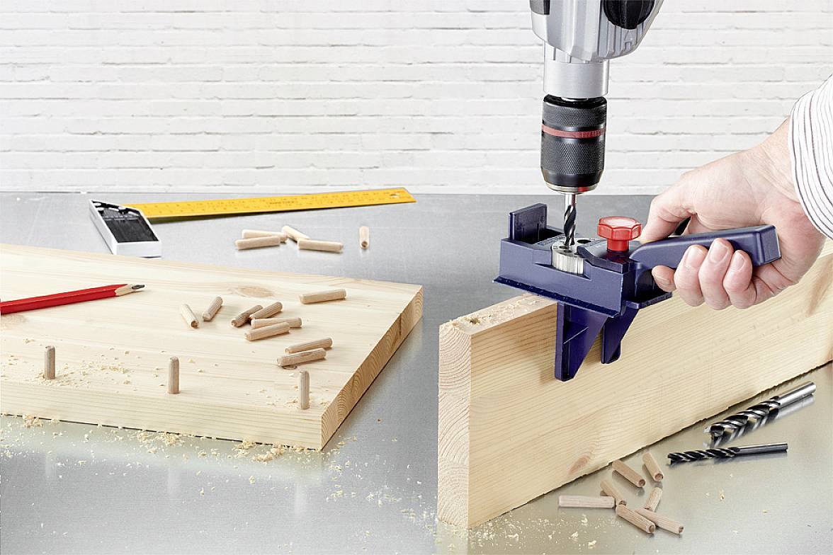 A craftsman is precisely drilling holes into a wooden board using a drill stand. Dowels and drill bits are lying on the table as working materials.