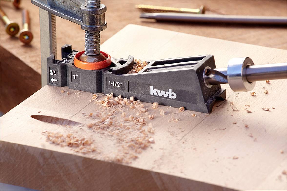 Drill template secured on wood, guiding the drill bit for precise drilling; wood shavings scattered around the hole. Work surface in the background.