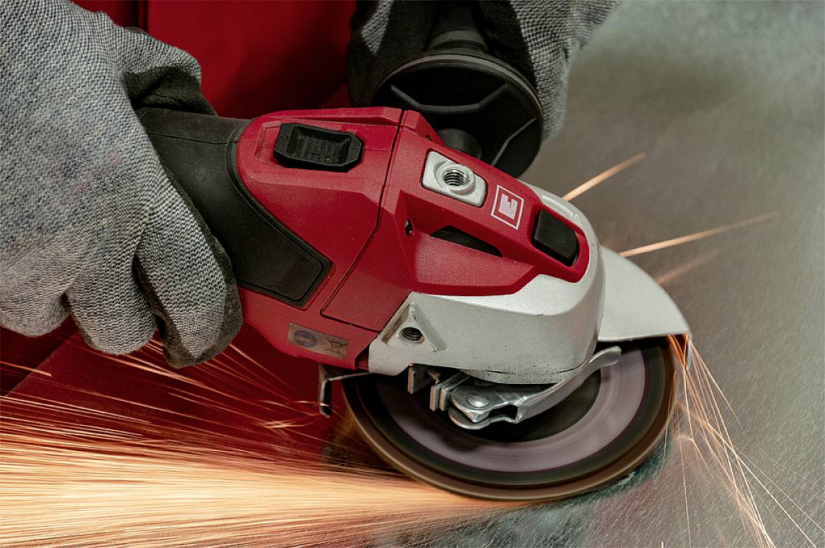 A person wearing grey gloves is using an angle grinder to cut metal, creating a substantial spray of sparks. Large sparks are clearly visible.