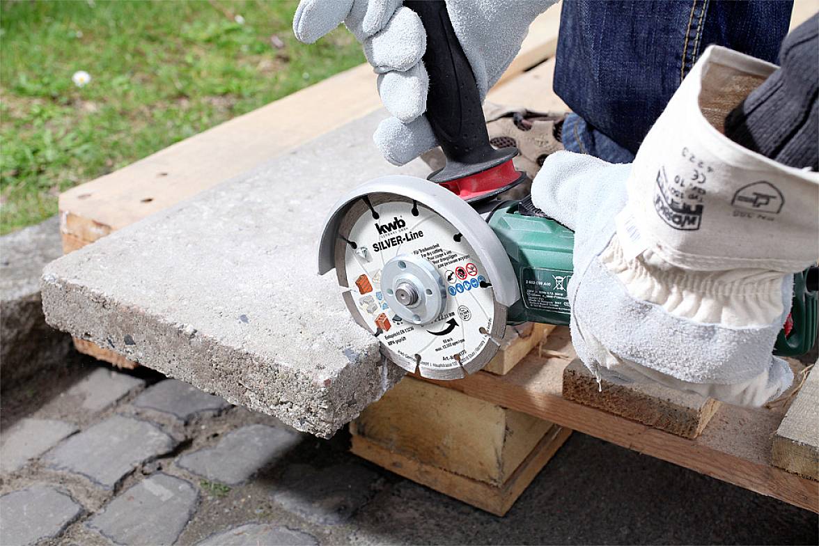 'Close-up of a person cutting a concrete block with an angle grinder. They are wearing gloves and standing on wooden planks.'