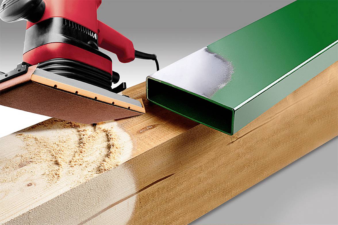 A red sanding machine smooths a wooden board. Beside it lies a green, rectangular metal profile on the board.