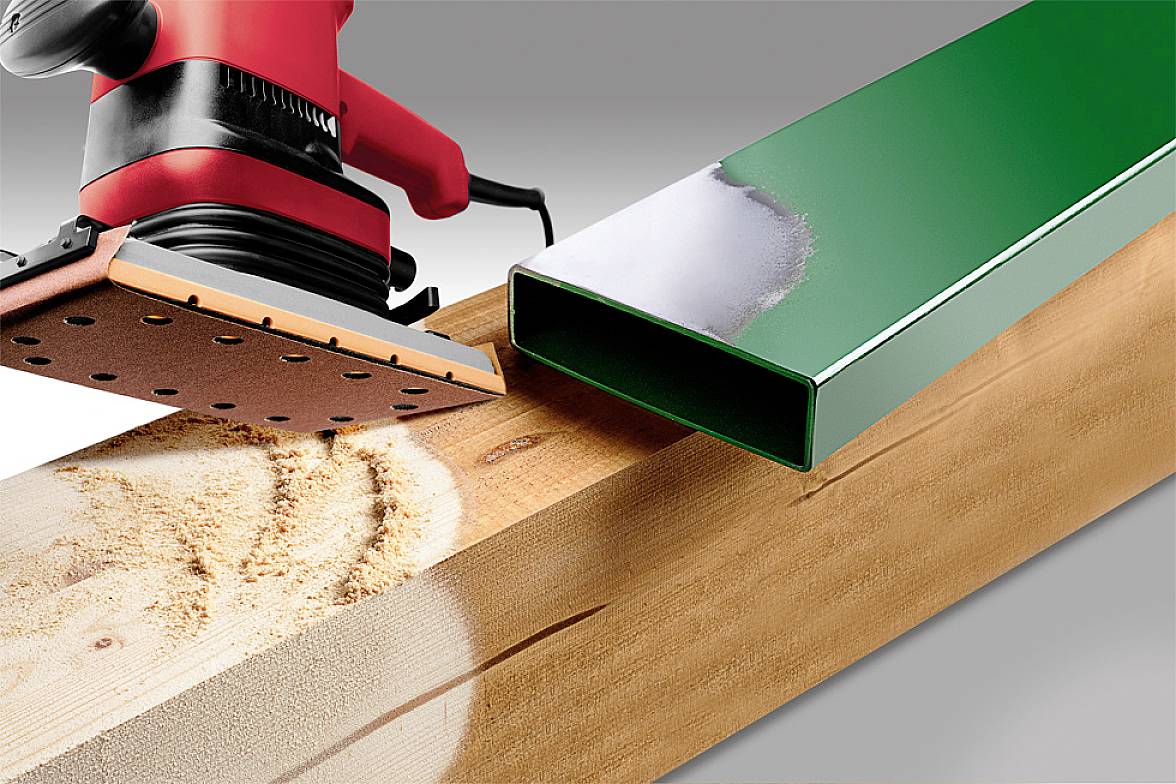 A red sander is working on a long wooden plank, with a green metal pipe lying beside it.