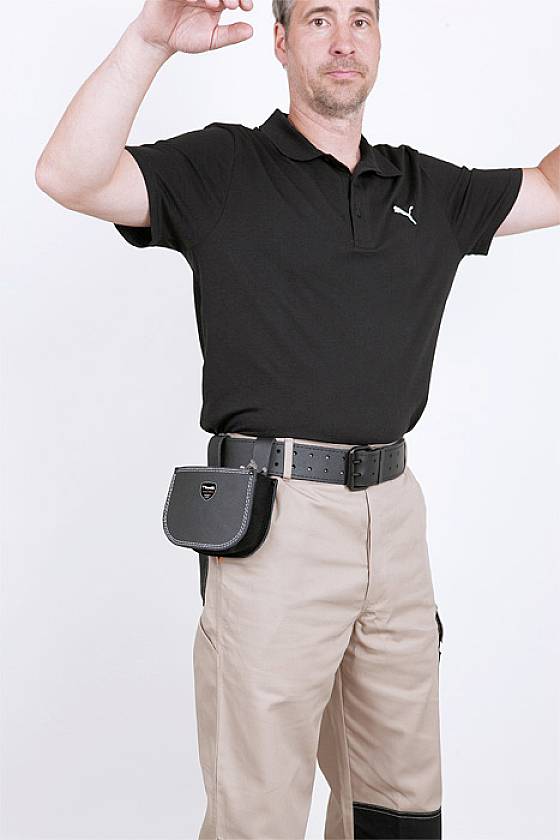 A person in a black polo shirt and beige trousers is raising their arms. They are wearing a black bag on a belt.