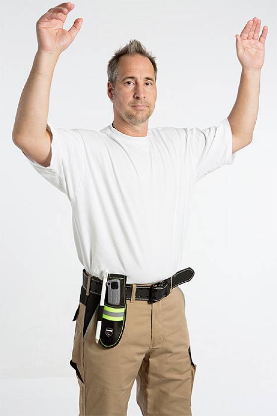 A man wearing beige trousers and a white T-shirt raises both arms. A device is attached to his belt. The background is white.