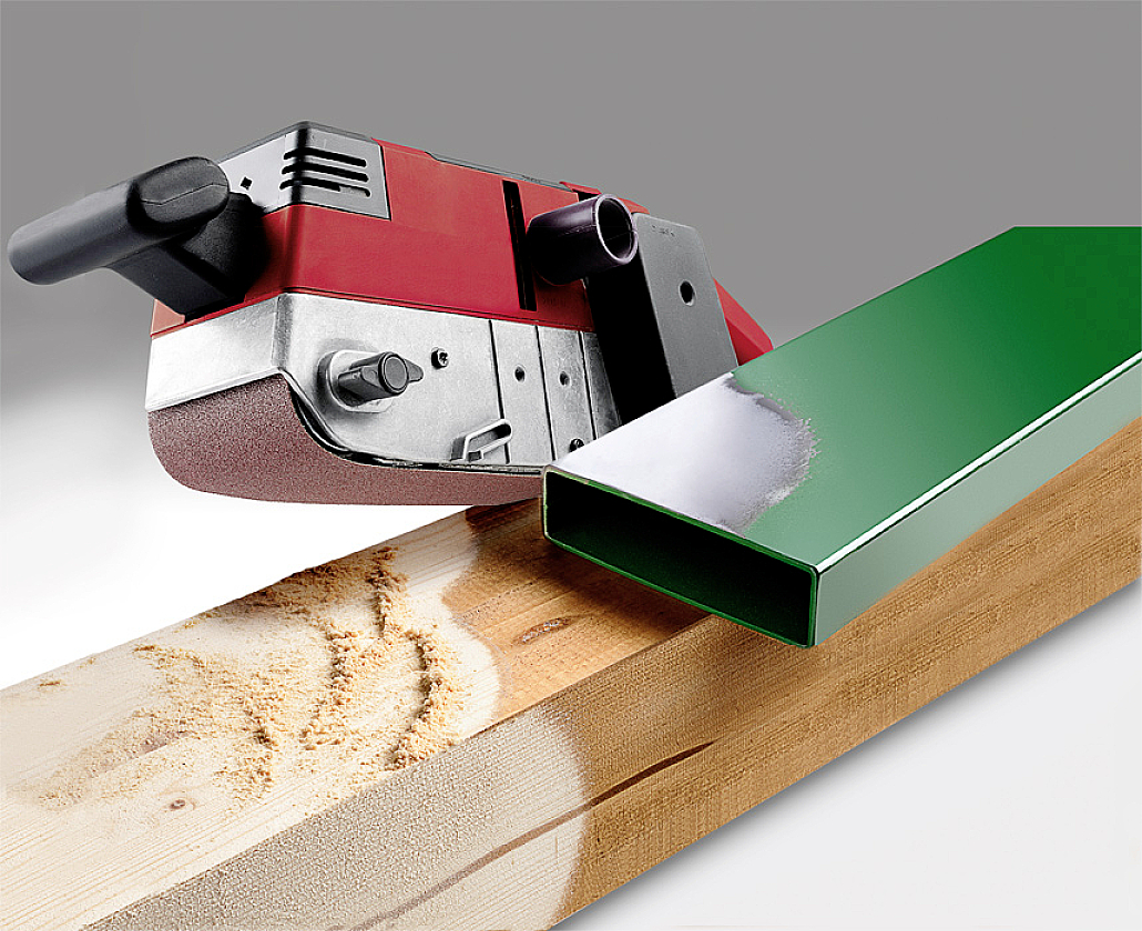 A red belt sander is working on a piece of wood, with a green rectangular metal tube lying beside it.