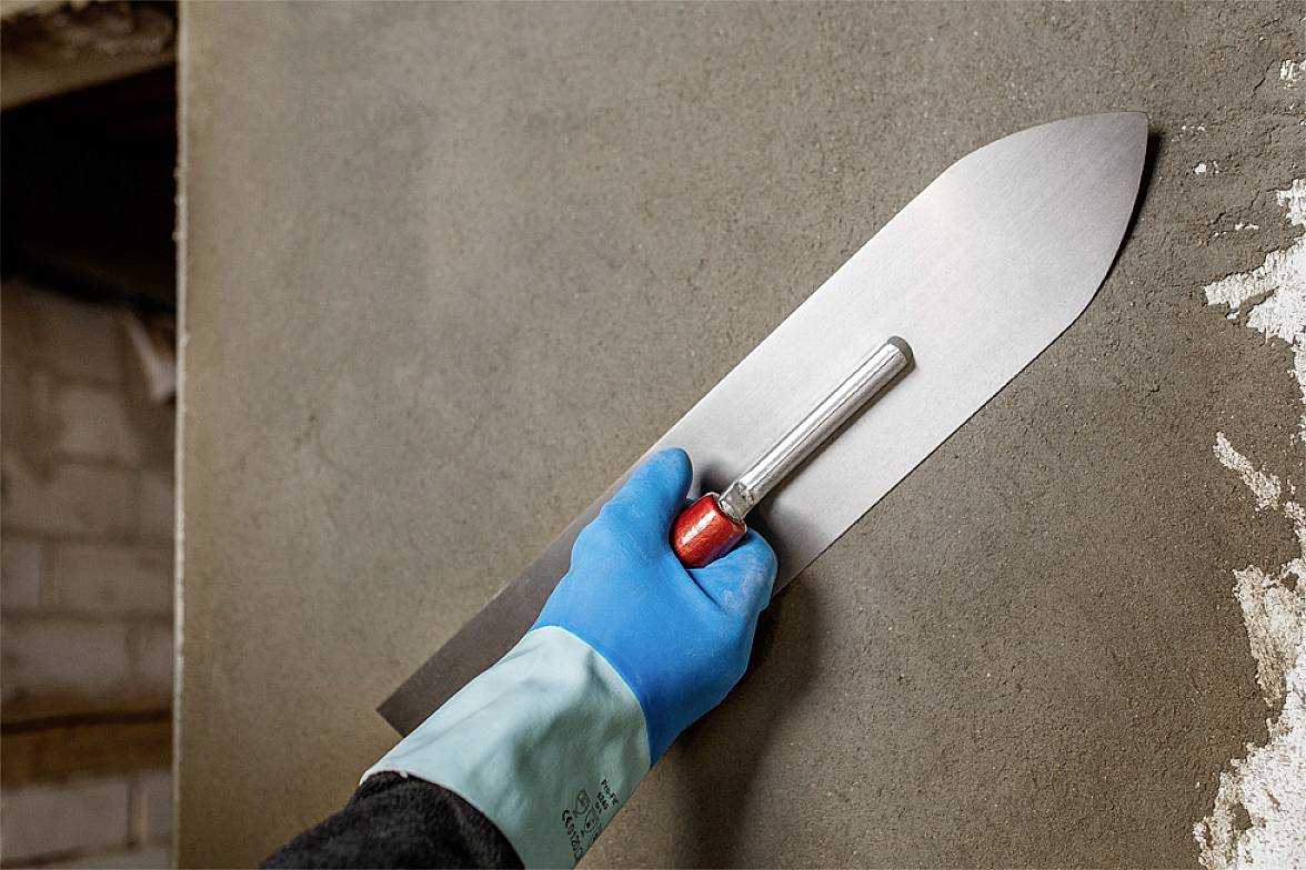 A person smooths freshly applied plaster on a wall using a trowel. They are wearing blue gloves for protection.
