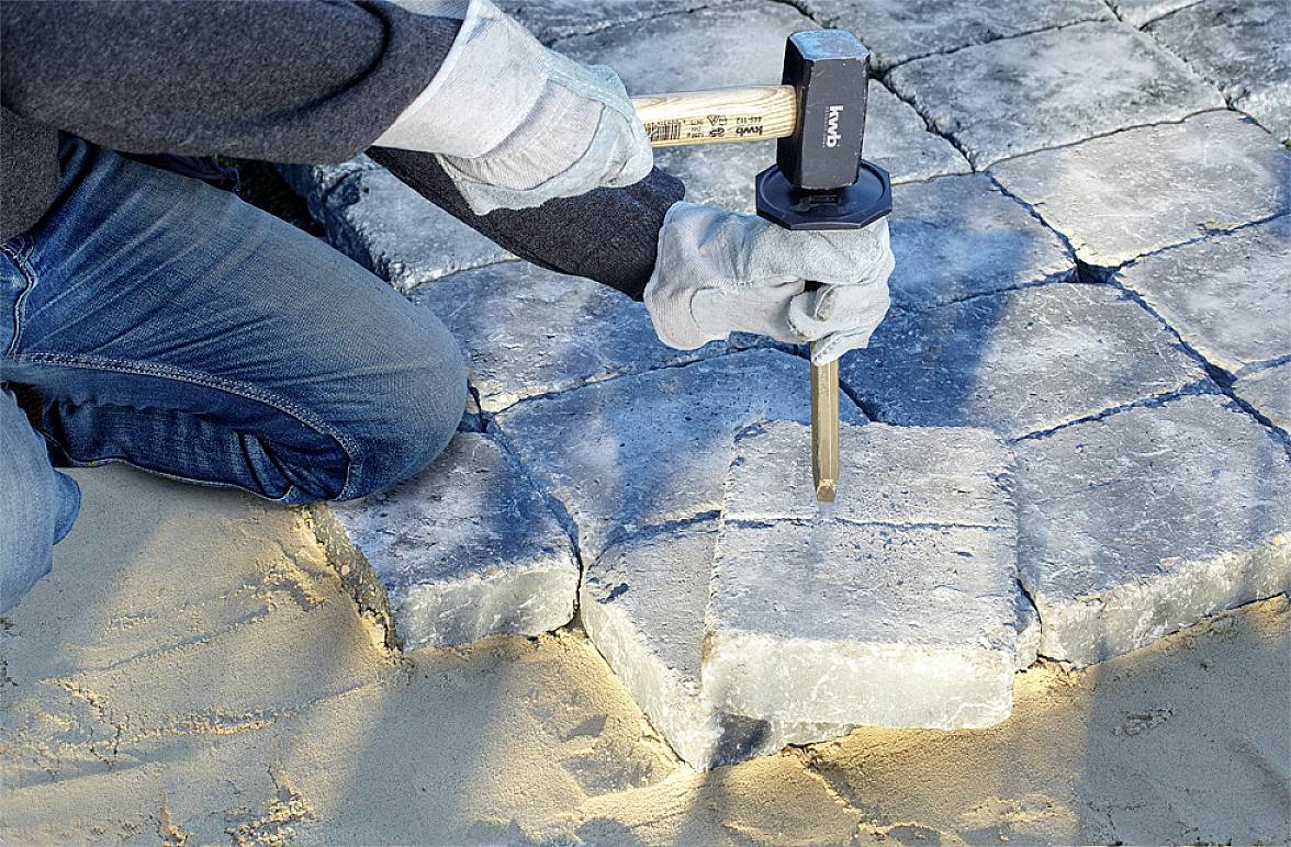 A person is laying stone slabs on a sandy surface and using a hammer and chisel to precisely fit the stones.