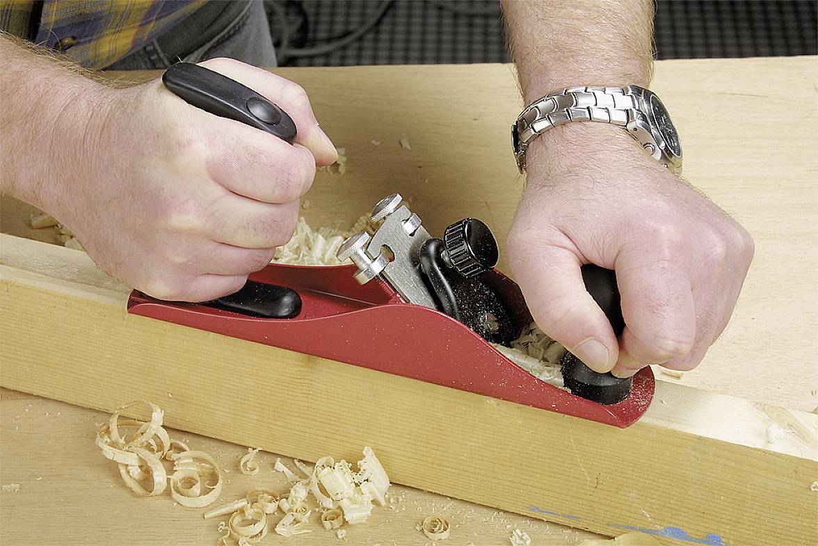 A person is planing a wooden board with a red hand plane. Wood shavings are visible on the table.